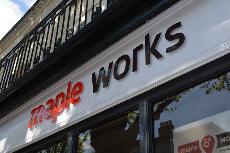 Close up of a of red and black letters that spell Maple Works. They're on a white decorative lintel above windows.